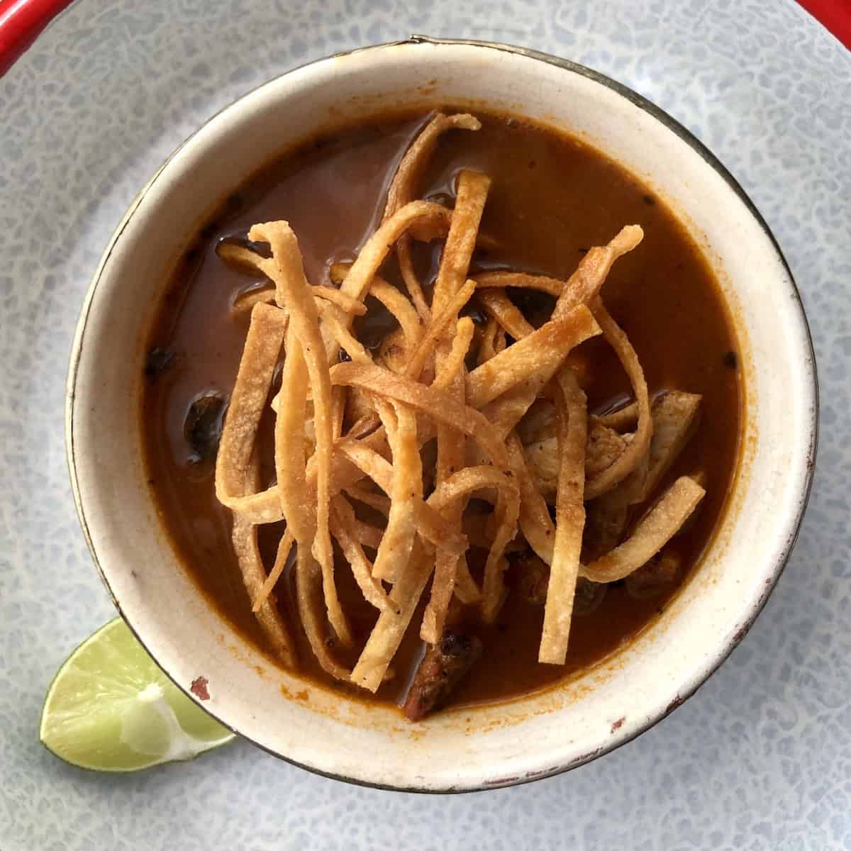 A full bowl of chicken tortilla soup with deep red broth, fried tortilla strips, pollo asado, avocado, crema, cheese, fried mulato chile, and a fresh lime wedge on the rim.