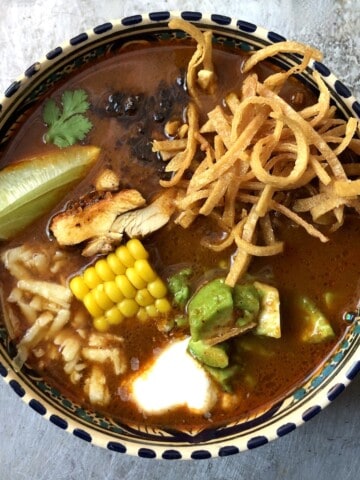 bowl of tortilla soup with crispy tortilla strips, pollo asado, corn, cheese, avocado, crema, fried mulato chile, a lime wedge, and a cilantro leaf.