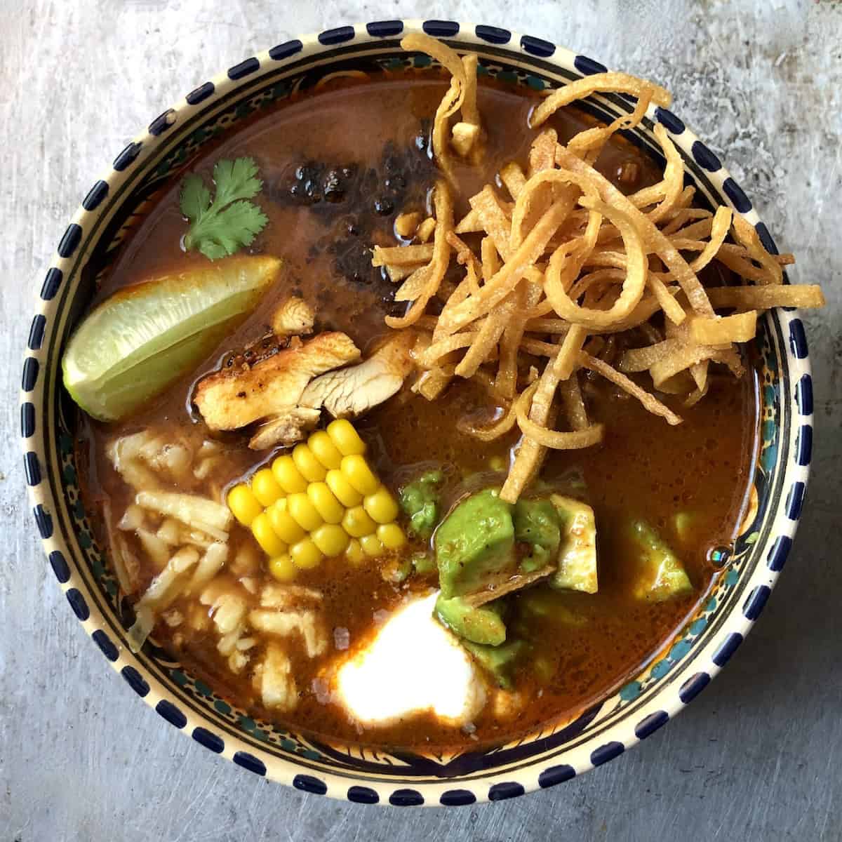 Bowl of chicken tortilla soup with crispy tortilla strips, pollo asado, corn, cheese, avocado, crema, fried mulato chile, a lime wedge, and a cilantro leaf.
