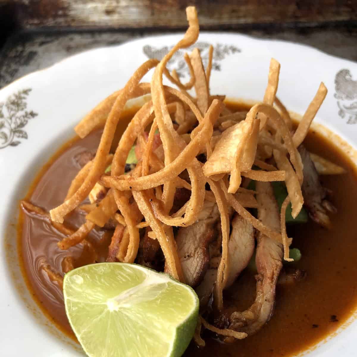 Closeup of chicken tortilla soup with crispy fried tortilla strips, sliced pollo asado chicken, avocado, and a lime wedge in a deep red chile and tomato chicken broth.
