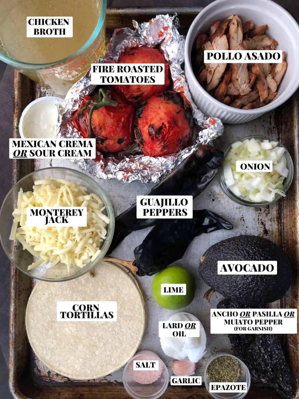 Labeled chicken tortilla soup ingredients on a sheet tray including cooked sliced pollo asado, homemade chicken broth, fire-roasted tomatoes, diced onions, guajillo chiles, mulato chile, lard, garlic, epazote, avocado, monterey jack cheese, corn tortillas, lime, Mexican crema, and salt.
