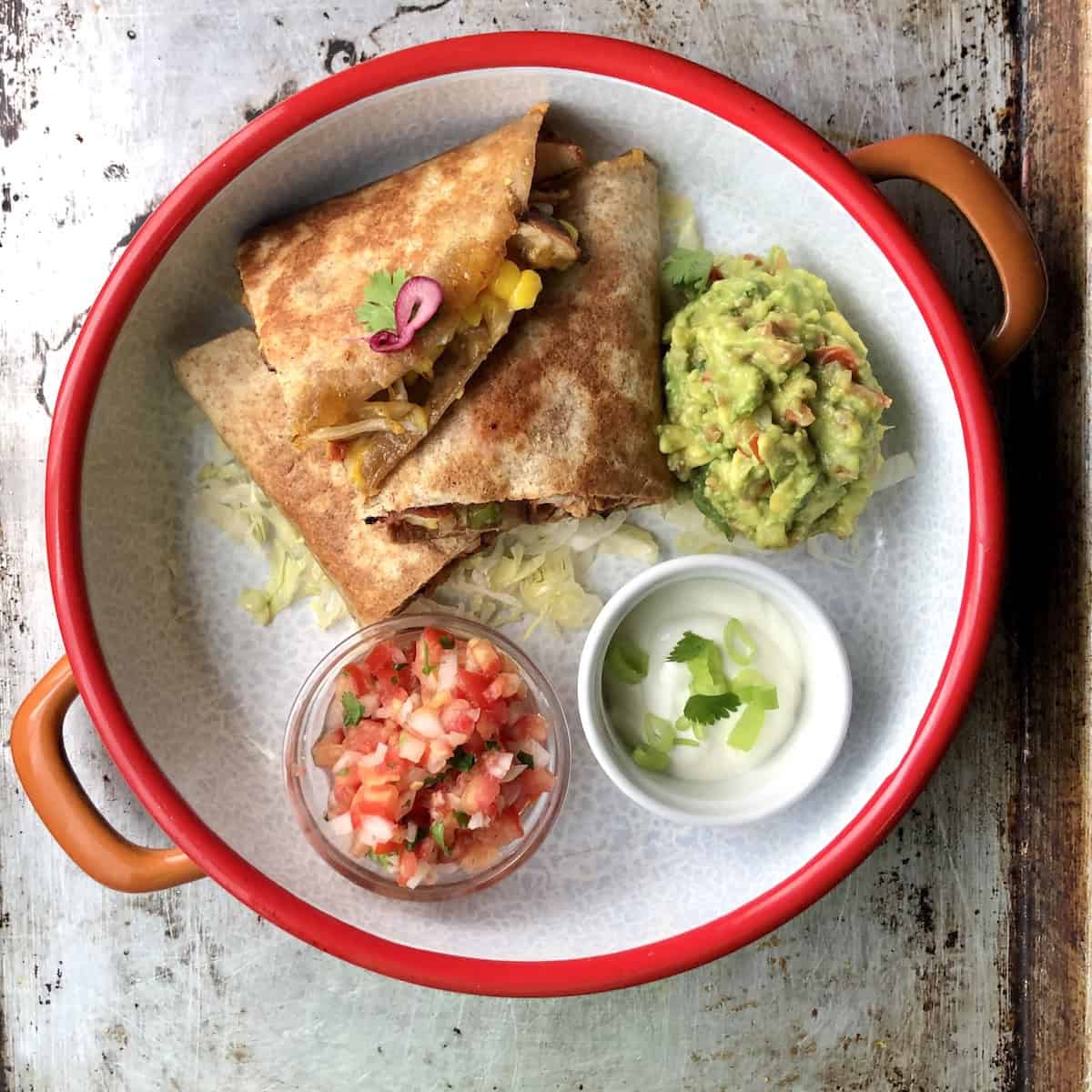 A fully cooked golden brown crispy chicken quesadilla on a serving platter with homemade salsa fresca, sour cream, and guacamole on a bed of shredded iceberg lettuce.