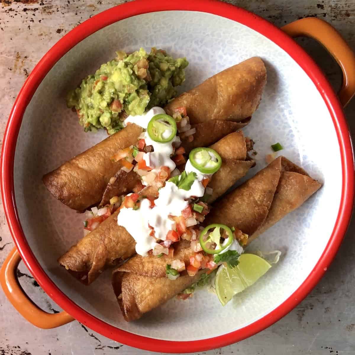 Three crispy flautas mexicanas on a colorful platter with shredded lettuce, homemade guacamole, salsa fresca, sour cream, fresh jalapeno slices, and a lime wedge.
