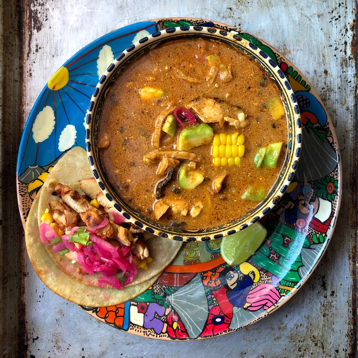 A bowl of creamy mexican tortilla soup stirred together with chicken, avocado, corn, and fried mulato chile next to a pollo asado taco with pickled red onions and cilantro on a colorful Mexican plate.
