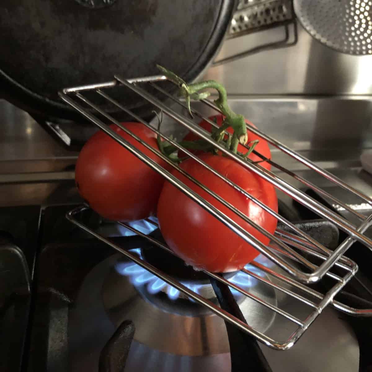 Three tomatoes with stems intact in a grilling cage set over a stovetop gas flame to fire roast for tortilla soup.
