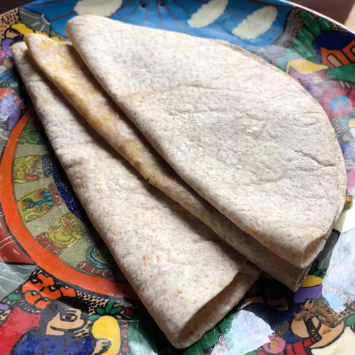 Folded whole wheat flour tortillas stacked on a colorful Mexican illustrated serving platter.


