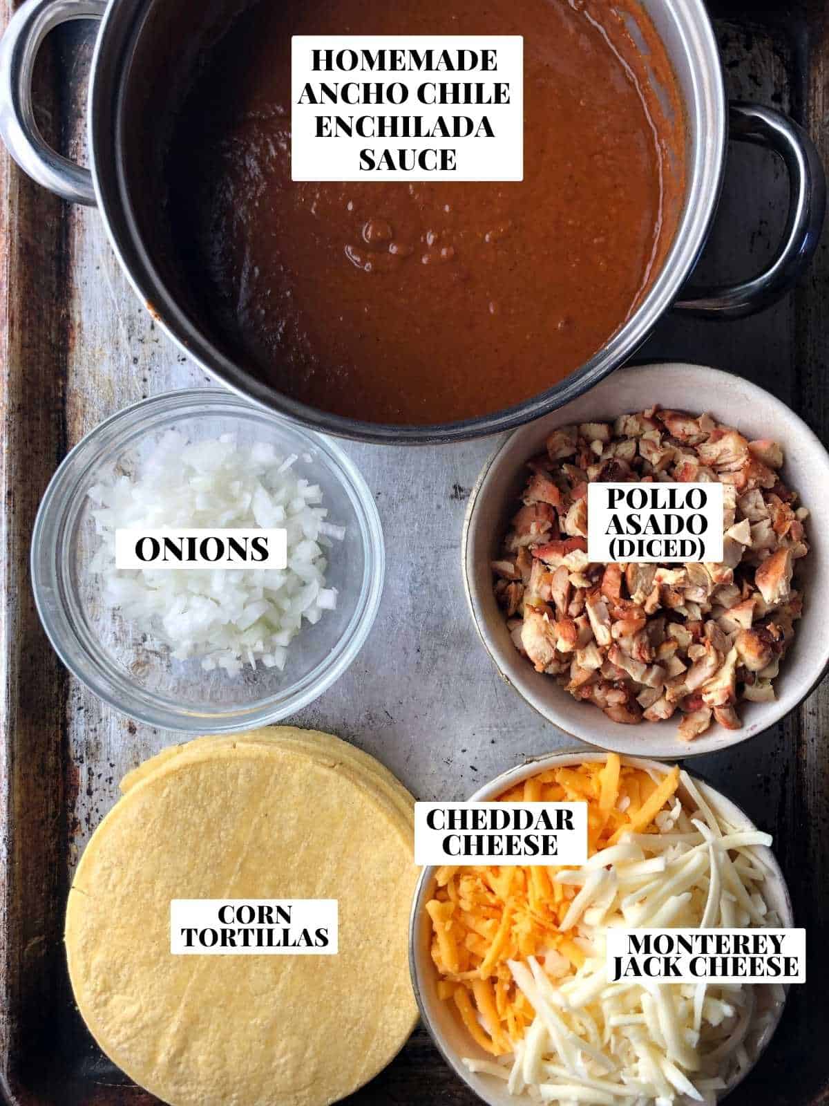 Labeled chicken enchilada ingredients on a sheet tray including diced pollo asado chicken, homemade ancho chile enchilada sauce, minced white onions, monterey jack cheese, cheddar cheese, and corn tortillas.