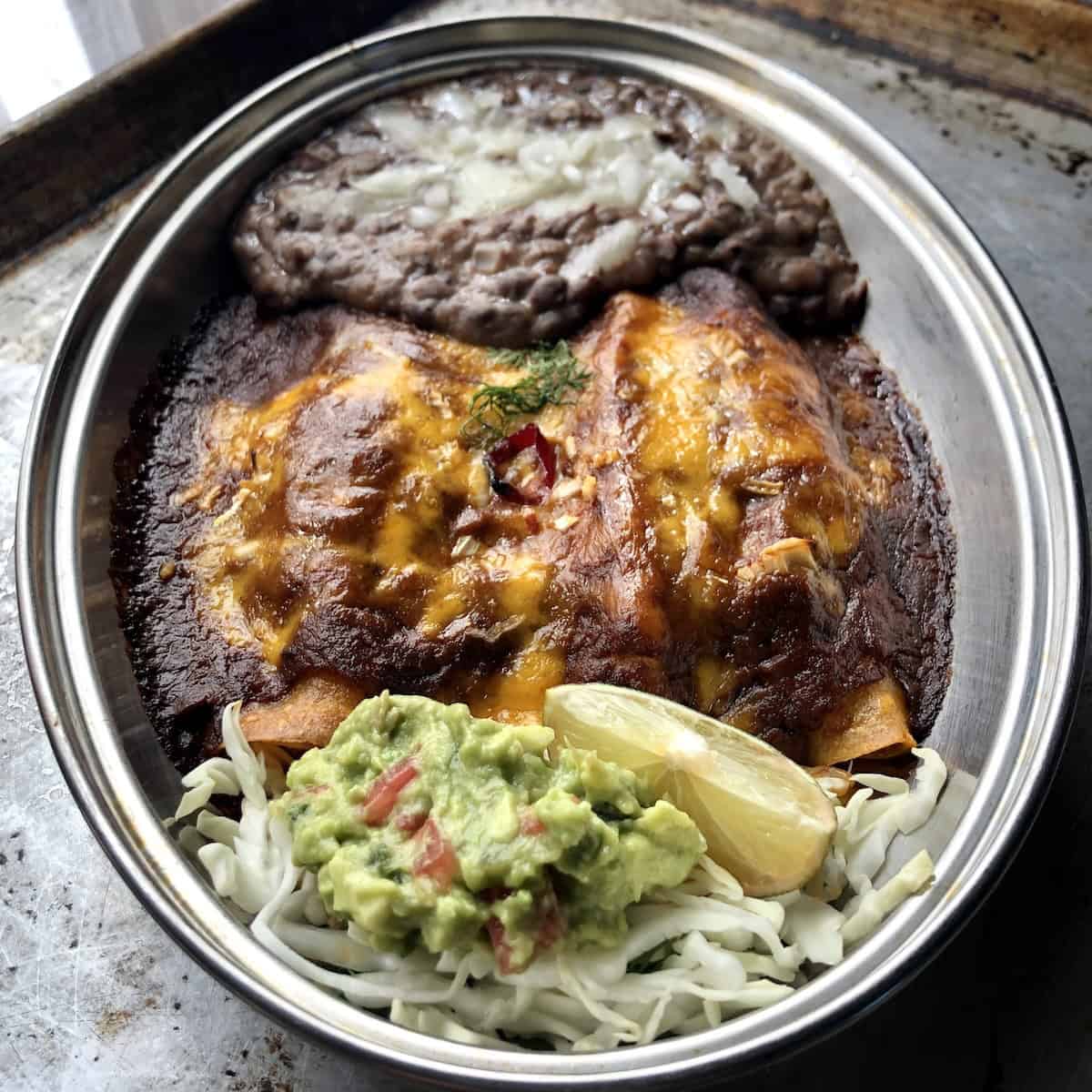 Three restaurant-style chicken enchiladas platter with homemade black refried beans, homemade guacamole, and a wedge of lime on a bed of shredded lettuce.