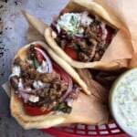 Easy gyro recipe with two pork gyros in a red basket served with a container of extra homemade tzatziki sauce on the side.