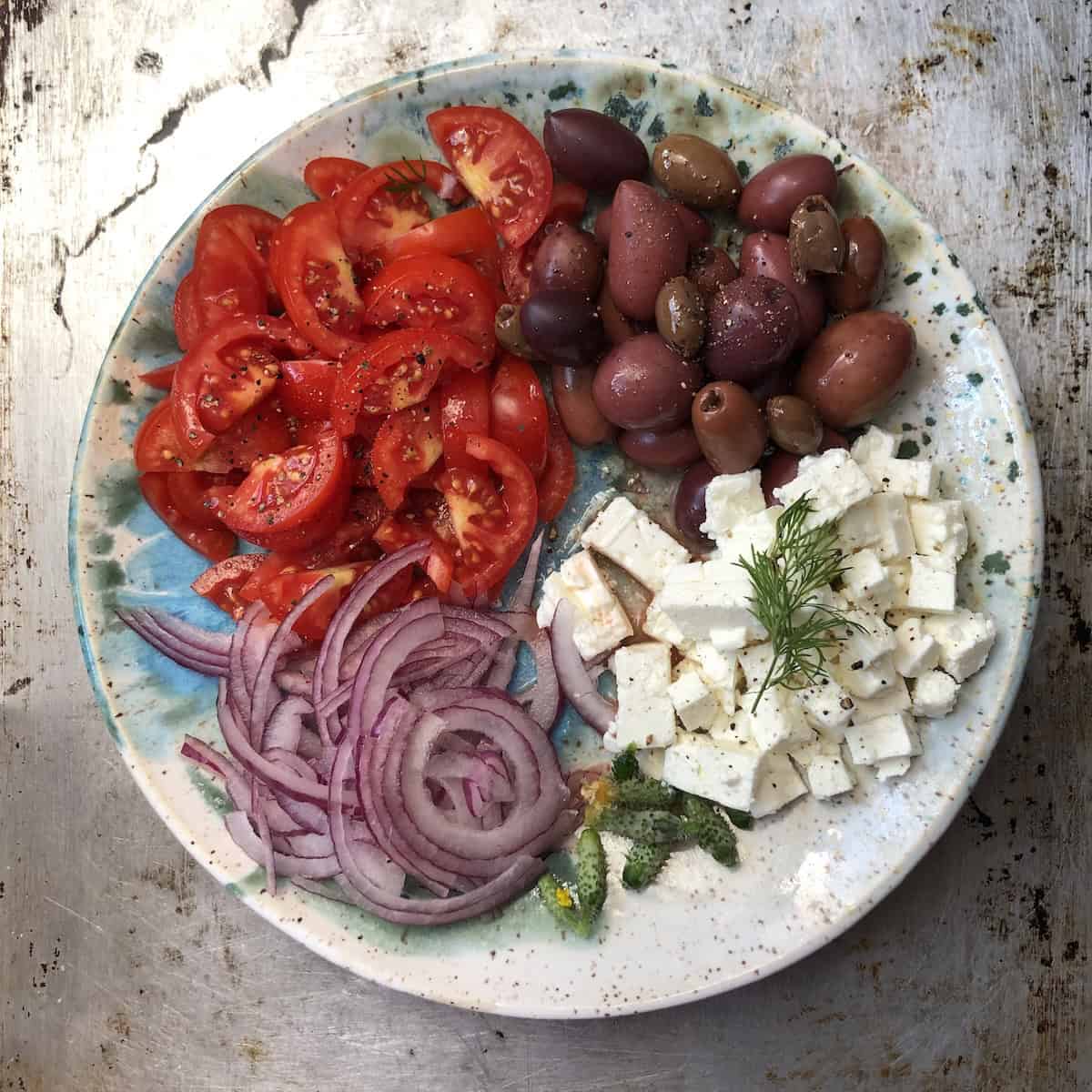Gyro toppings on a plate including Greek olives, feta cheese cubes, fresh dill, sliced cucumbers, grape tomatoes, and red onions for a Greek gyro sandwich.
