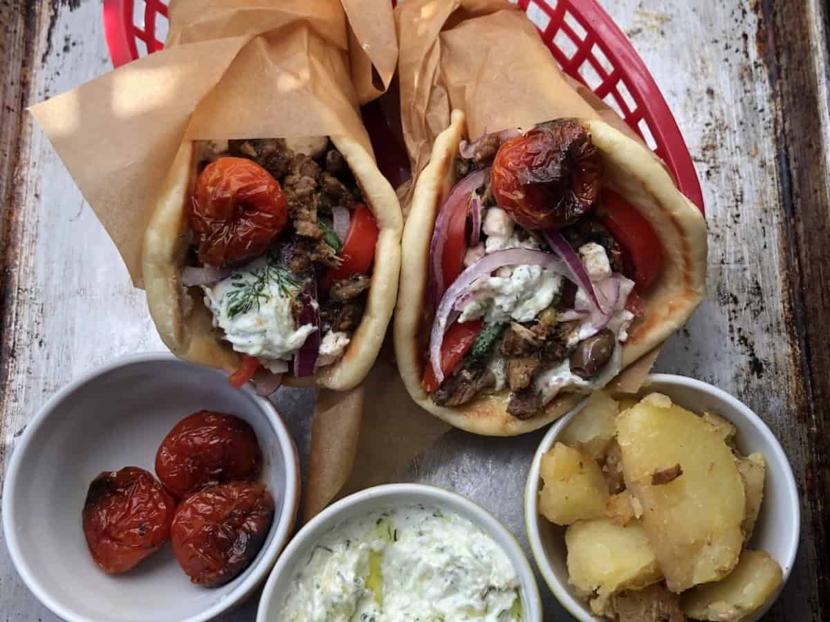 Homemade gyro recipe closeup of a pork gyro sandwich in a red basket with bowls of roasted tomatoes, tzatziki sauce, and roasted gyro potatoes.