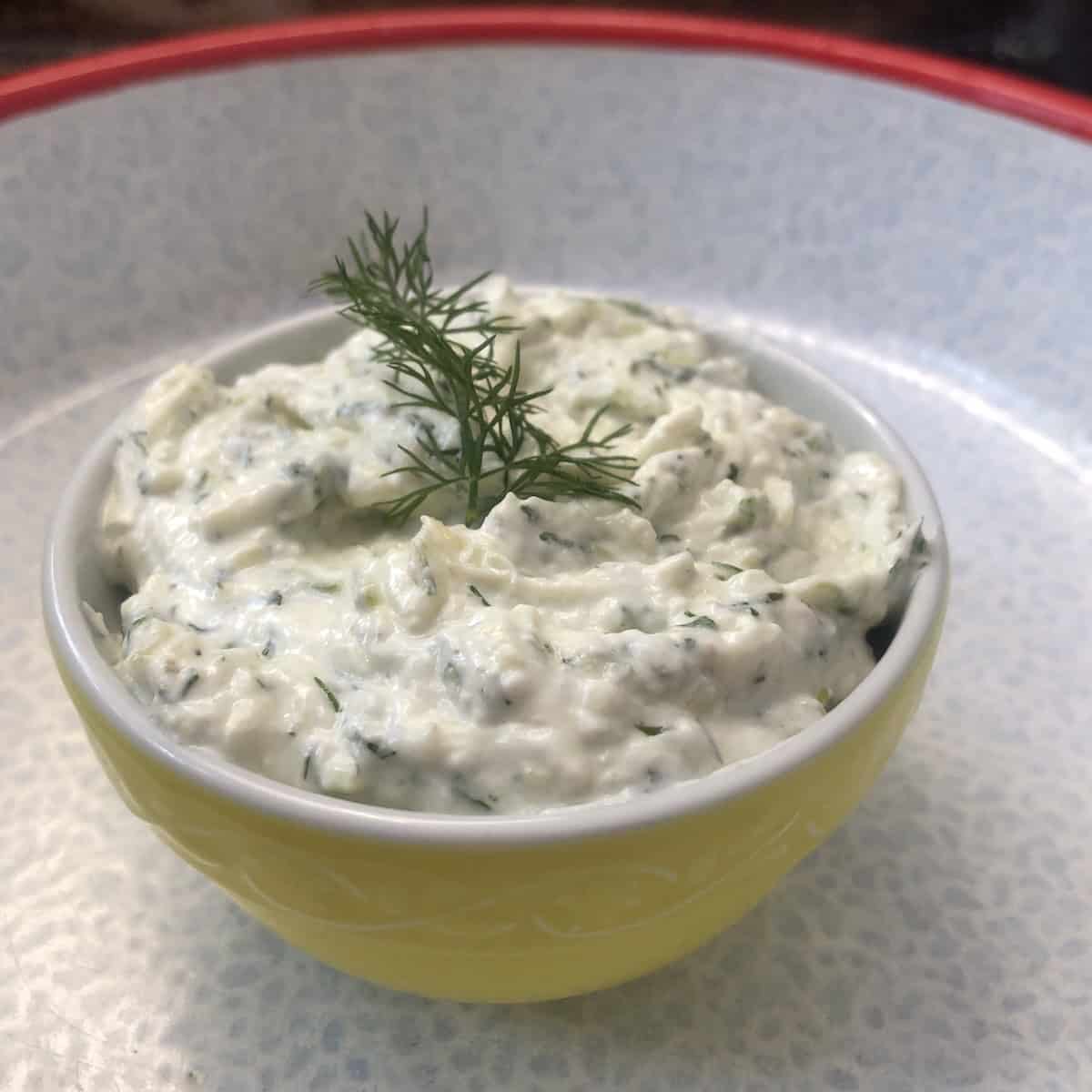 Homemade tzatziki sauce in a yellow bowl topped with a sprig of fresh dill.