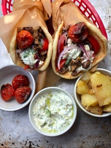 Two Greek gyro sandwiches in red baskets made with homemade greek pita and tzatziki sauce, gyros meat, feta cheese, olives, tomatoes, cucumbers, and red onions, with bowls of roasted tomatoes, extra tzatziki, and roasted gyro potatoes in front.