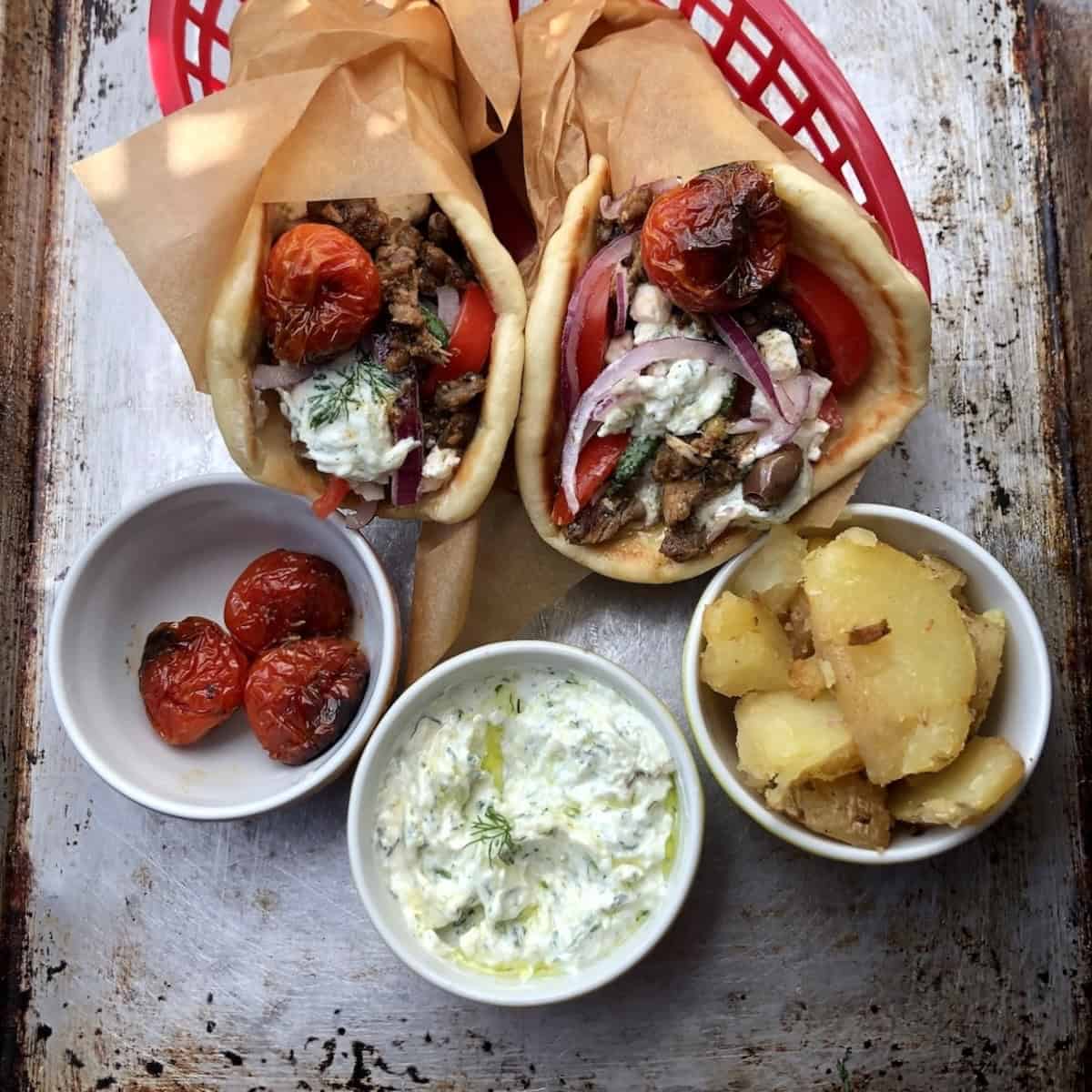 Two Greek gyro sandwiches in red baskets made with homemade greek pita and tzatziki sauce, gyros meat, feta cheese, olives, tomatoes, cucumbers, and red onions, with bowls of roasted tomatoes, extra tzatziki, and roasted gyro potatoes in front.
